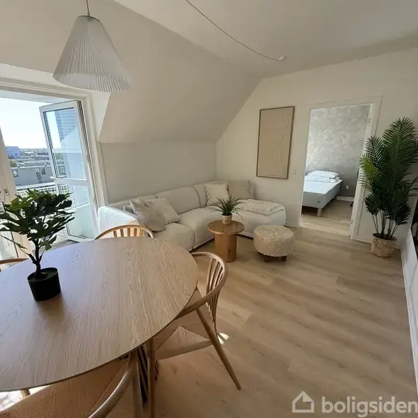 En lys stue indeholder en hjørnesofa med puder og planter på et rundt sofabord. Et spisebord med stole står ved siden af en åben altandør. En åben dør fører til soveværelset.