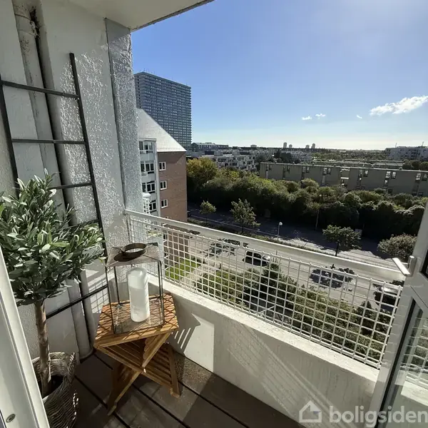En altan med træbord og lanterne står stille, omgivet af grønne planter. Udsigt over en byvej med biler og træer, og en blå himmel i baggrunden.