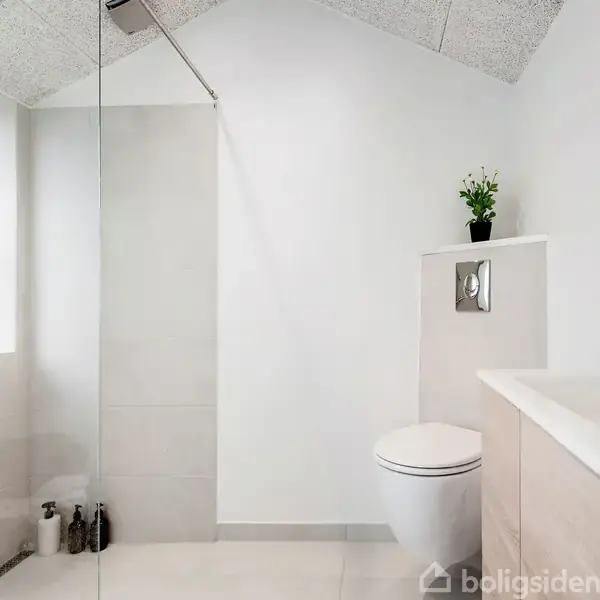 Et lyst badeværelse med et bruseområde til venstre, en toilet med en lille plante ovenpå og en vask med spejl til højre. Rummet har moderne indretning.