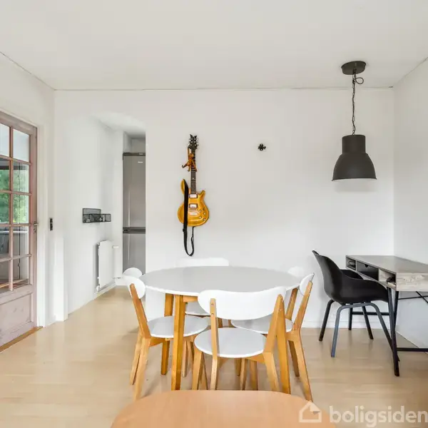 Rundt bord med fire stole står i en lys stue. På væggen hænger en guitar. Ved siden af er et skrivebord med en sort stol og en stor lampe.