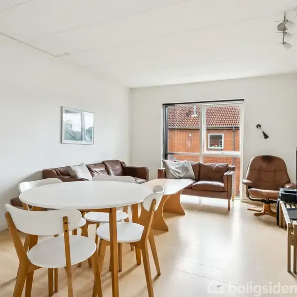 En stue med et hvidt spisebord og stole i forgrunden. To sofaer og en lænestol er placeret ved et stort vindue, suppleret med en lampe og fjernsyn.