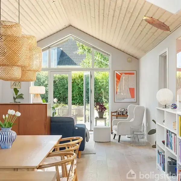 Spisebordet står centralt med træstole og vase med blomster. Store lamper hænger over bordet. I baggrunden ses en opholdsstue med sofa, reol og store vinduer mod haven.