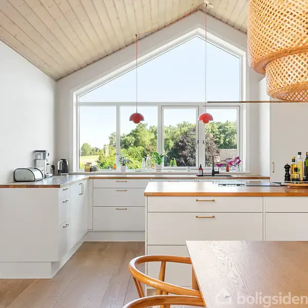 Køkken med hvidt inventar arrangeret i en L-form. Et stort vindue med udsigt til en grøn have. En flettet lampe hænger over et træbord med tulipaner.