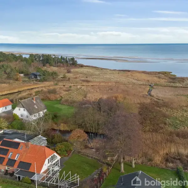 Flere huse med røde tage står stille nær en kyst. Omkring dem er der grønne marker og en kystlinje med siv, mens havet strækker sig mod horisonten under en blå himmel.