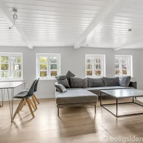 Grå sofa står i en lys stue med trægulv og hvide vægge. Rummet har fire små vinduer og et sofabord. Der står også et spisebord med to sorte stole.