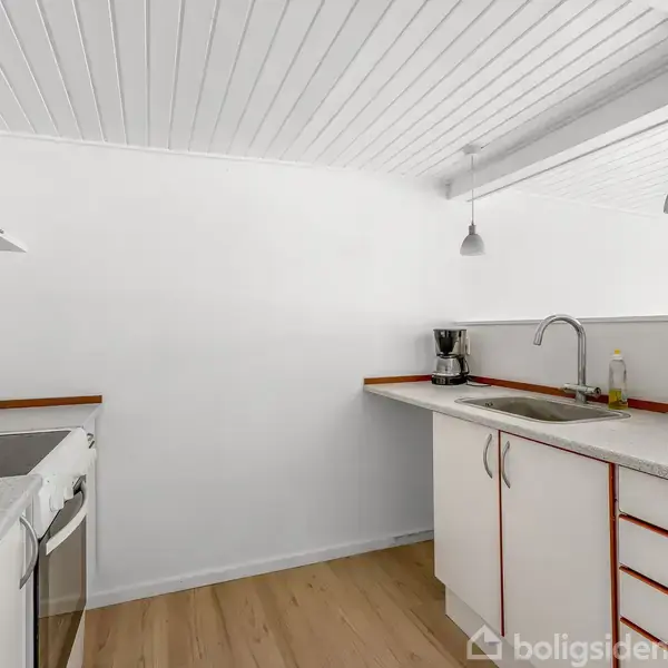 Køkken med hvide skabe og bordplader, en indbygget ovn og kogeplader til venstre, kaffemaskine ved siden af vasken, og to hængende lamper i loftet, omgivet af hvide vægge.