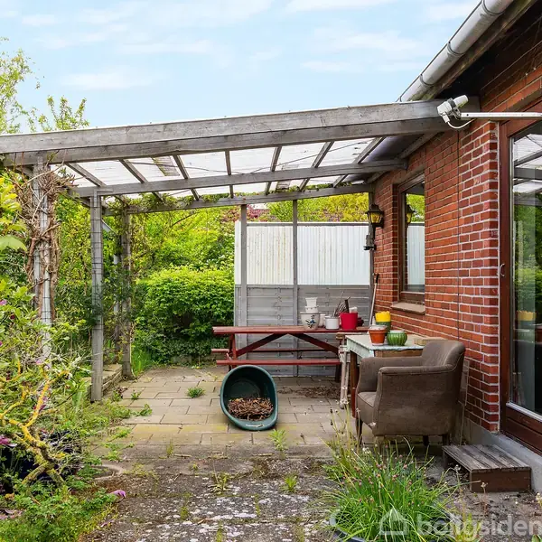En terrasse med en sofagruppe og bord sidder under en pergola ved et murstenshus. Omkring er der blomster og buske, og i baggrunden står en rød bænk.
