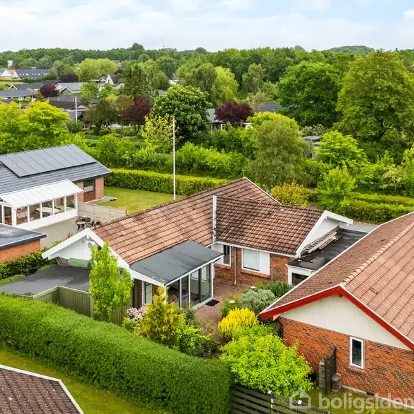 Huset har rødt tag og er omgivet af grønne hække. Det ligger i et villakvarter med flere lignende huse. Baggrunden består af træer og en blå himmel.