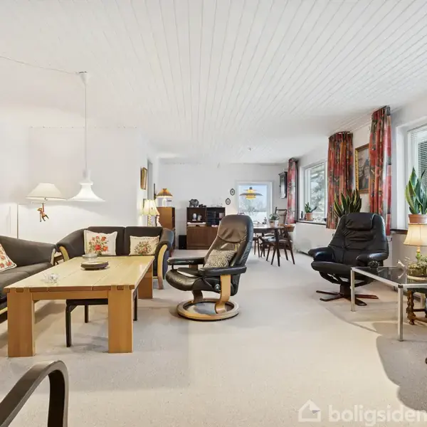 Stue med sorte sofaer og lænestole arrangeret omkring et sofabord, dekoreret med planter og lamper. Store vinduer med røde gardiner lukker lys ind, og et maleri hænger på væggen.