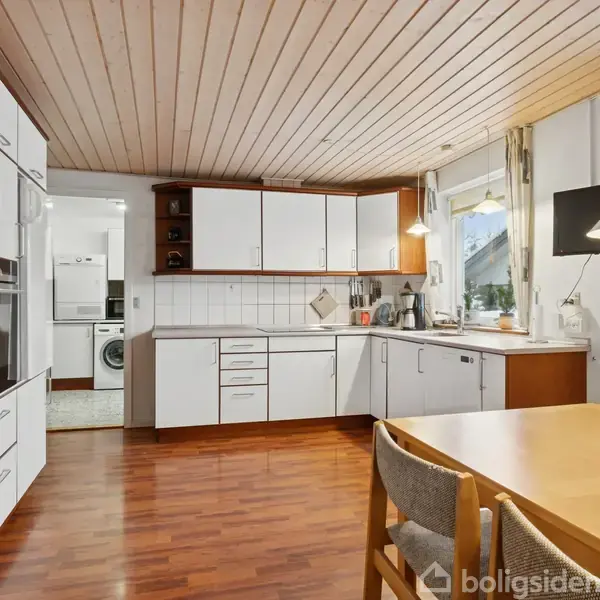 Et køkken møbleret med hvide skabe, en bordplade og indbyggede apparater. Et træbord med stole står i forgrunden. Rummet har trægulv og lyst loft. Vinduet har udsigt til haven.