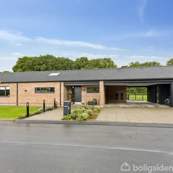 Et moderne enfamiliehus med fladt tag står i en åben have. Det har et træforet område på siden og en carport. Grønne buske og træer omkranser ejendommen.