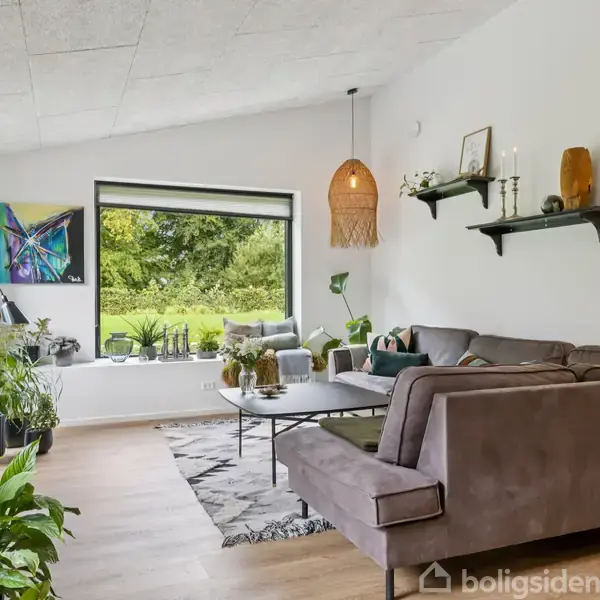 En hjørnesofa står i en stue med lyse vægge. Et stort vindue viser en grøn udsigt. Planter og kunst dekorerer rummet, der har hylder med dekorationer og en lampeskærm.