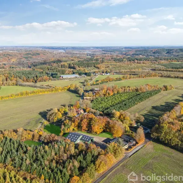 Luftfoto viser en gård med solpaneler omgivet af marker og skov i efterårsfarver, der skaber en varieret og farverig landskabsudsigt under en delvist skyet himmel.