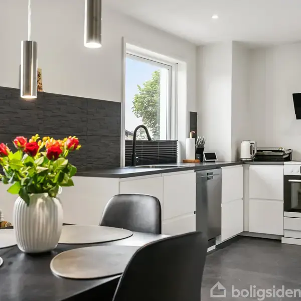 Spisebord med blomsterbuket i vase står i moderne køkken. I baggrunden er der et komfur, opvaskemaskine, ovn og sorte køkkenskabe. Vinduer giver naturligt lys.