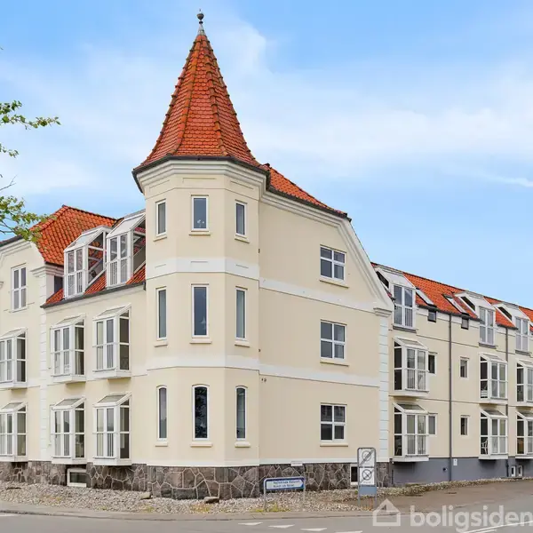 En hjørnebygning med hvid facade og rødt tegltag, inklusive et tårn, står langs en gade. Blå himmel og træer omgiver bygningen.