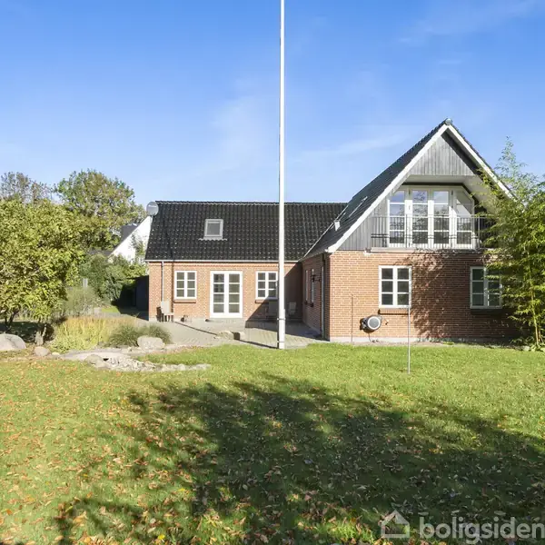 Rødt murstenshus med hvide vinduer står stille i en grøn have med græs og træer. En flagstang er placeret foran huset under en klar blå himmel.