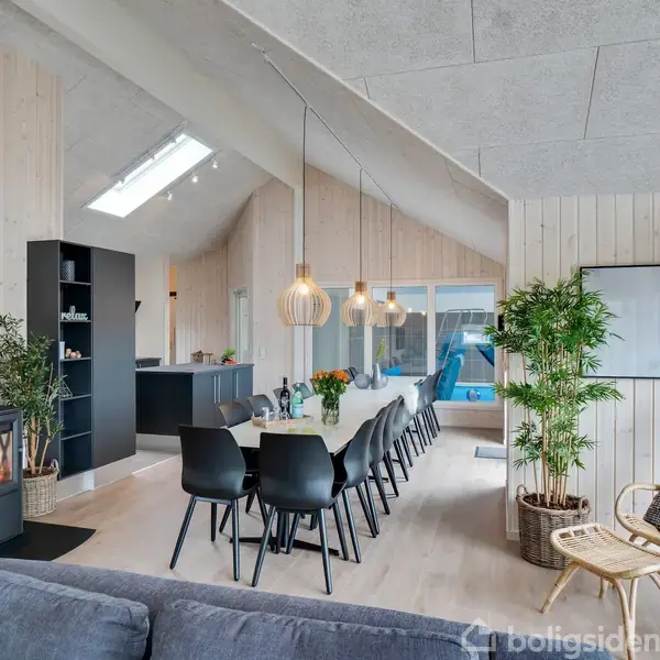 Langt spisebord med sorte stole står i en moderne stue. Over bordet hænger to dekorative lamper. I baggrunden ses en grå sofa og planter foran store vinduer.