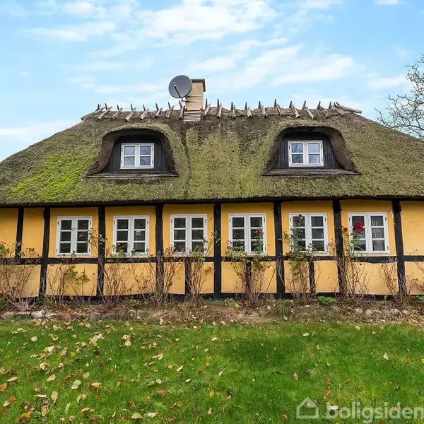 Et gult bindingsværkshus med stråtag står i en grøn have. Seks små vinduer pryder forsiden, omgivet af en træindhegning. Et satellitfad sidder på taget.