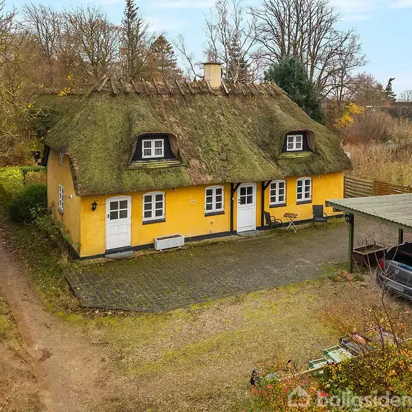 Gult hus med stråtag, står stille på en stor grund omgivet af træer. En bilparkeret under carport til højre, grusvej til venstre fører væk fra huset.