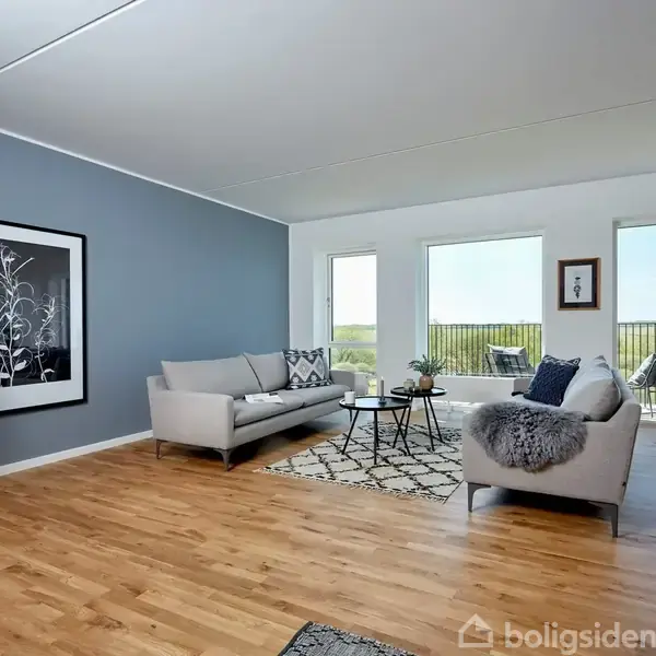 En stue med to lysegrå sofaer placeret omkring et sofabord, planter og vægkunst. Rummet har trægulv, en stor blå-grå væg og store vinduer med udsigt til en altan.