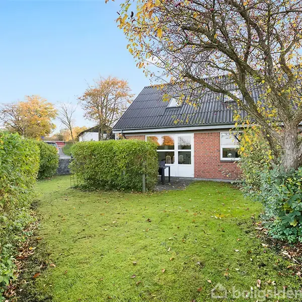 Hus med røde mursten og sort tag ses i baggrunden. Det står omgivet af en velplejet have med grøn græsplæne og høje buske. En stor træ står i nærheden.