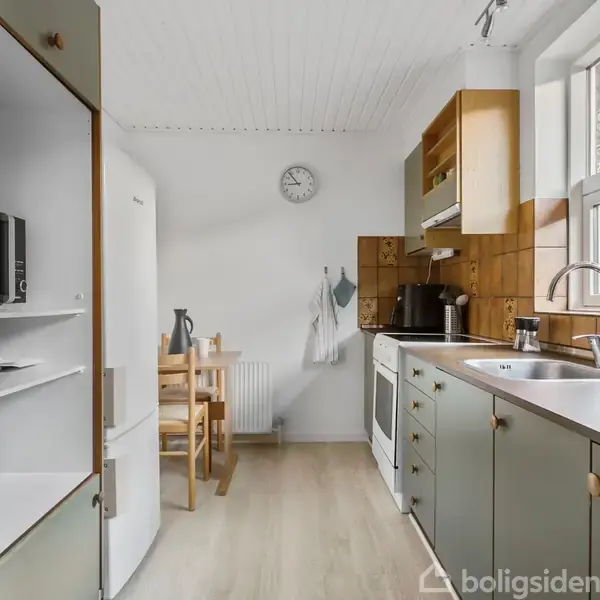 Køkken med hvid køleskab og mikrobølgeovn til venstre. Komfur og vask til højre under vinduer. Træbord og stole i baggrunden. Ur hænger på væggen.