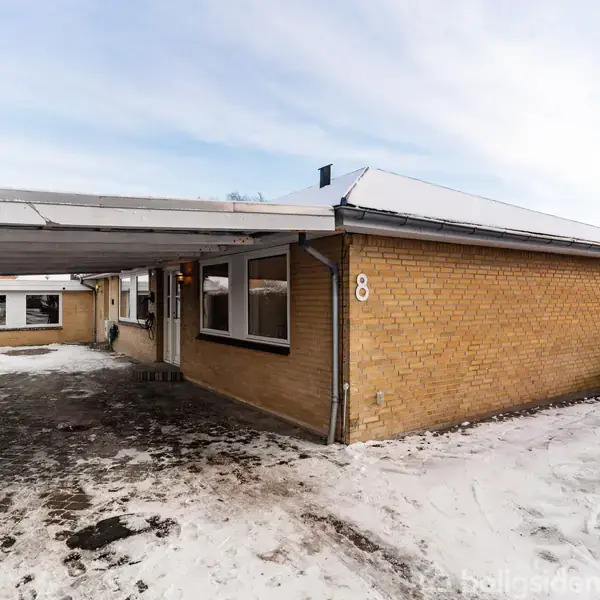 En gul murstensbygning med carport står i et snedækket område. Tallet "80" er synligt på væggen nær indgangen.