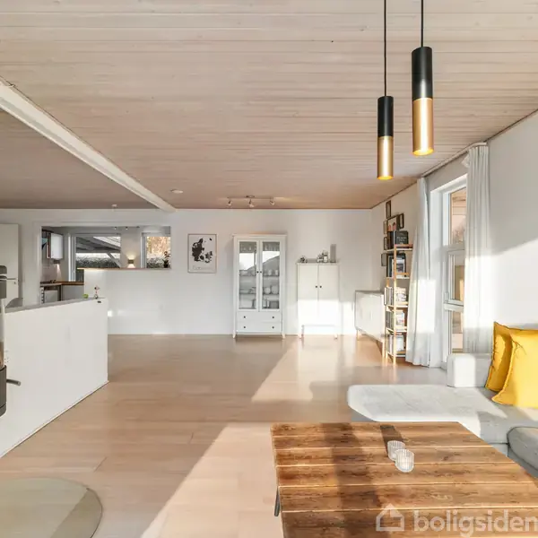 En moderne stue med en brændeovn til venstre. En grå sofa med gule puder står ved et stort vindue. Hængelamper hænger fra loftet. En træbord står i midten.