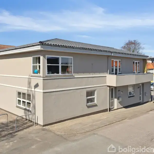 En tofamiliers ejendom står på et hjørne i et byområde. Bygningen er to etager høj med beige mure og store vinduer. Parkeringsplads og beboelseshuse ses i baggrunden.