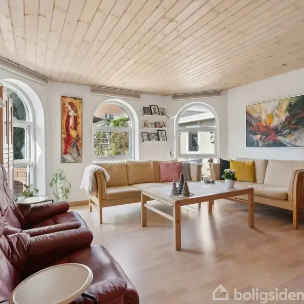 Røde lænestole og en beige sofa står i en stue med træloft og store buede vinduer. Væggene er dekoreret med farverige malerier. Et sofabord står i midten.