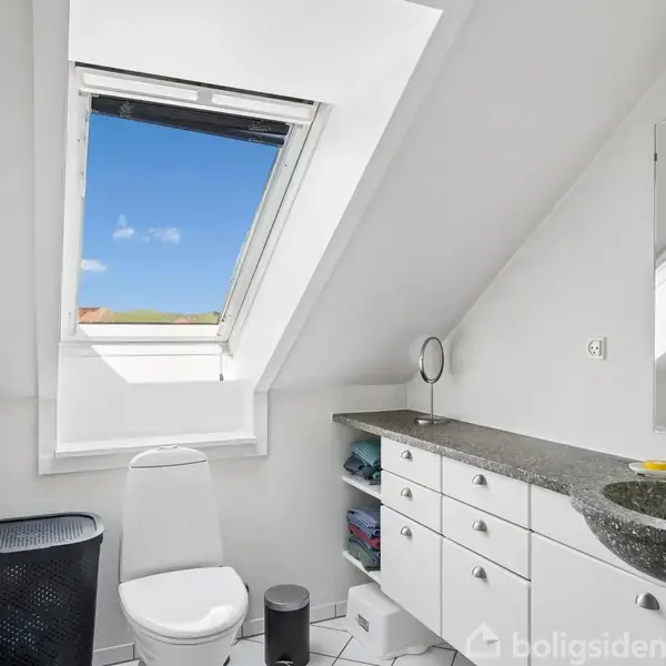 Et lyst badeværelse med toilet under et åbent skråvindue. Håndvasken er placeret på en sort bordplade med hvide skabe nedenunder. En rund spejl ses på bordet.