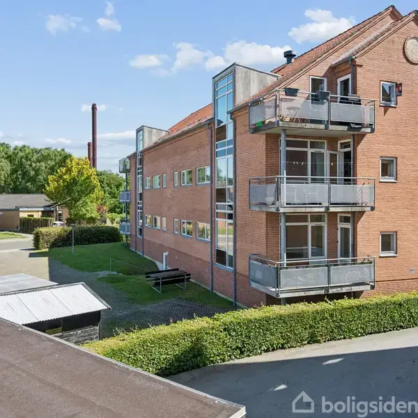 En rød murstensejendom med altaner strækker sig op mod en blå himmel. Omgivelserne består af grønne træer og buske, med en vej og et andet hus i nærheden.