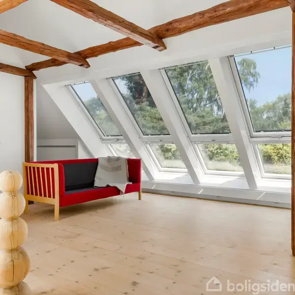 Rød sofa står stille i et lyst loftsrum med synlige træbjælker og store skråvinduer, der giver udsigt til træer udenfor.