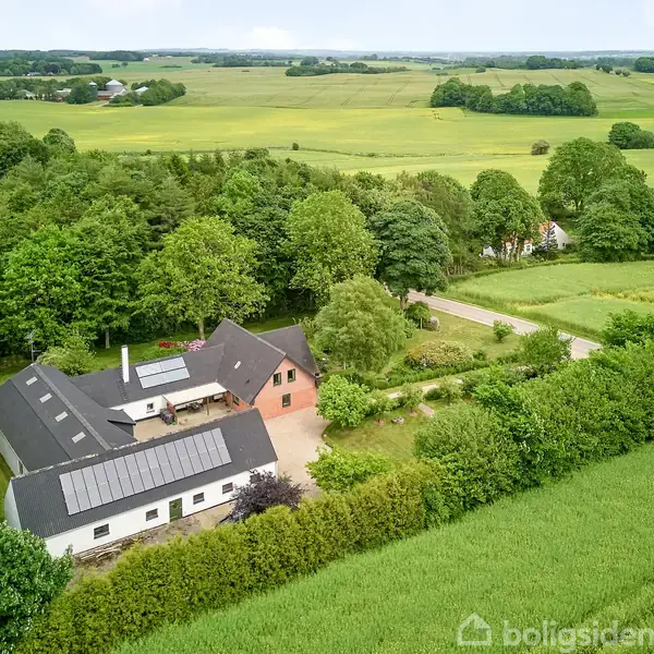 Et større landhus med solpaneler på taget ligger omgivet af frodig vegetation og marker. En landevej snor sig forbi ejendommen i et grønt, landligt område.