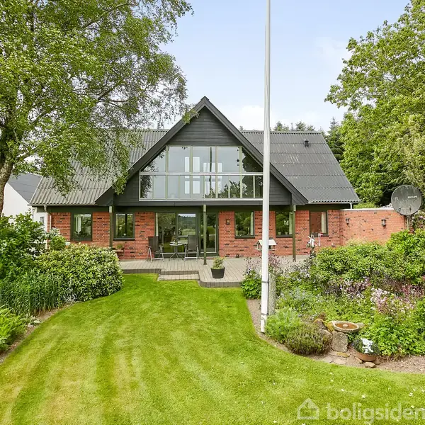 Huset har en sort tagkonstruktion og store glaspartier. Det står i en have med grønne plæner og diverse planter, omgivet af træer. Der er en flagstang foran huset.