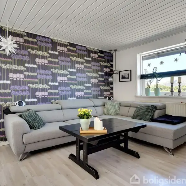 Grå sofa med grønne puder står i en stue. Et sofabord har planter på toppen. Baggrunden har mørkt tapet med blomstermønster og en rektangulær vindue. Ingen tekst til stede.