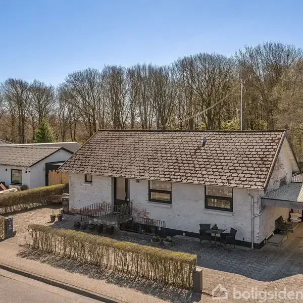 Et hvidt stenhus med sadeltag står stille på en stille villavej, omgivet af en velplejet hæk og parkeringsplads. I baggrunden ses en tæt skov med høje træer.