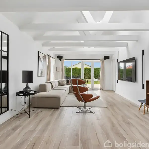 En moderne stue med hvide vægge og træloft. Sofagruppe står ved store vinduer, der fører ud til en have. På væggen er der også et stort spejl og en sort lampe.