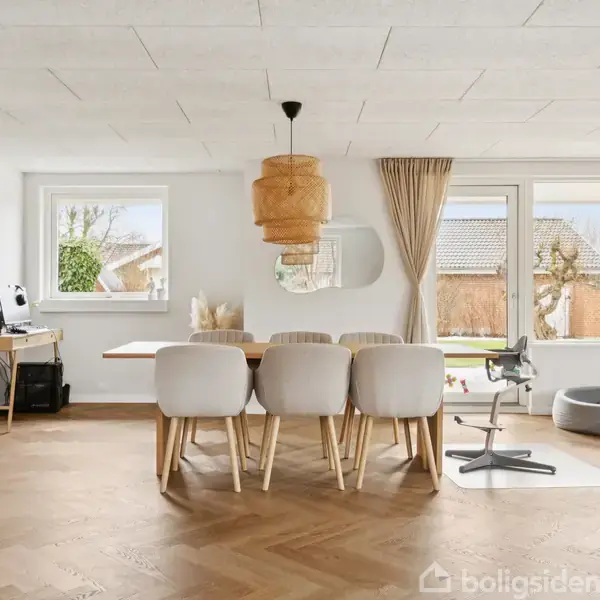 Spisebord med seks stole i lys stue, omgivet af store vinduer med udsigt til have. Trægulv, loftlampeskærm og skrivebord med kaffemaskine til venstre.