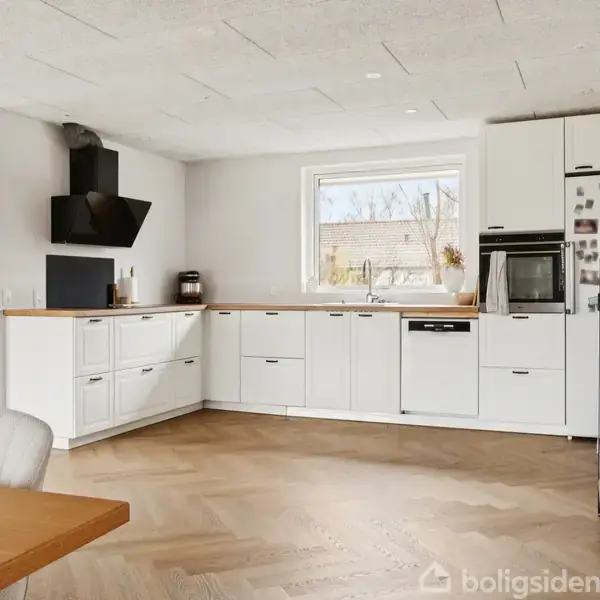 Et moderne køkken med hvide skabe, en sort emhætte og træbordplader, står i et rummeligt værelse. Et vindue giver udsigt udenfor. En glasvitrine indeholder glas. En sofa er synlig i baggrunden.