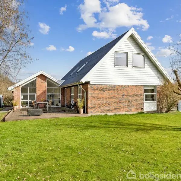 En rødmuret villa med hvide detaljer står på en grøn græsplæne. Terrasse med havemøbler ses foran huset. Træer omgiver ejendommen under en blå himmel med få skyer.