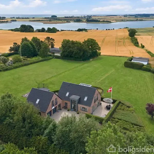 Et stort hus med en mørk tag ligger på en velholdt græsplæne. Området er omgivet af marker og træer med en fjord i baggrunden. En dansk flag står udenfor.