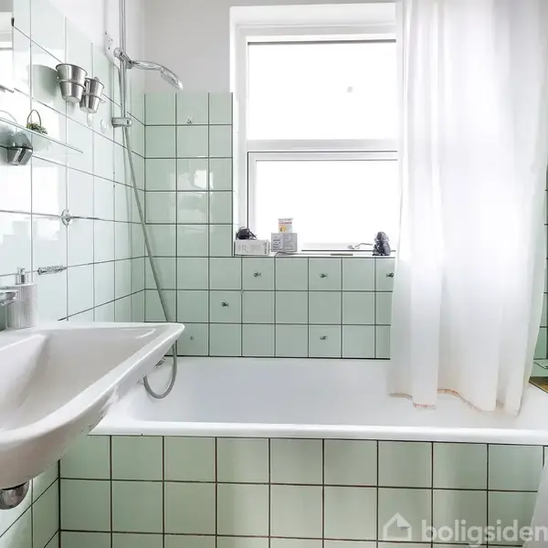 Badekar med brusehoved og hvidt badeforhæng. Vask monteret til venstre. Lysegrønne fliser dækker vægge og kar, lille vindue i baggrunden. Hylde over vasken.