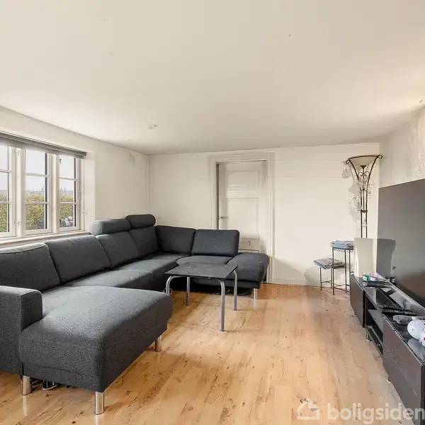 En stor sort sofa står i en stue og danner en hjørnevinkel. Foran står et lavt bord. Til højre findes et fjernsyn på et mediemøbel. Rummet har trægulv og hvide vægge.