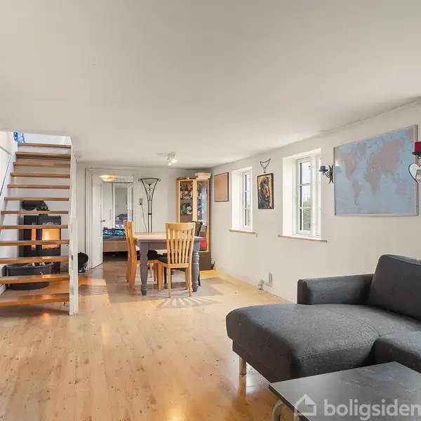 Sofa i gråt stof står i en stue med trægulv, ved siden af et spisebord med stole. Trappen i baggrunden leder til en øvre etage, omgivet af hvide vægge.