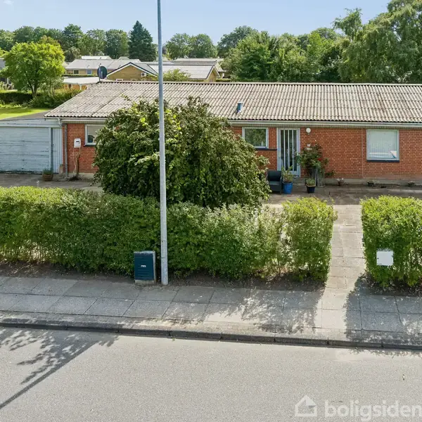 Rødt murstenshus med fladt tag og stor forhave med grøn hæk, garage til venstre. Omgivet af andre huse i et grønt boligområde.