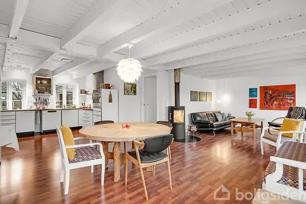 Et lyst, åbent rum med træmøbler og planter ved skråvinduer. I baggrunden ses en græsplæne gennem store glasdøre, som lader dagslys komme ind i rummet.