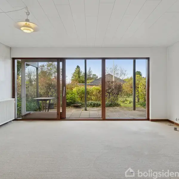 En stue med tæppebelagt gulv har store glasdøre, der åbner ud til en terrasse. Udenfor er der udsigt til en grøn have. Rummet er lyst med en enkelt lampe.