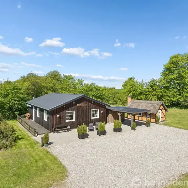 Et sort træhus og en mindre bindingsværkbygning står på en grusbelagt plads omgivet af grønne træer og marker under en blå himmel med spredte skyer.