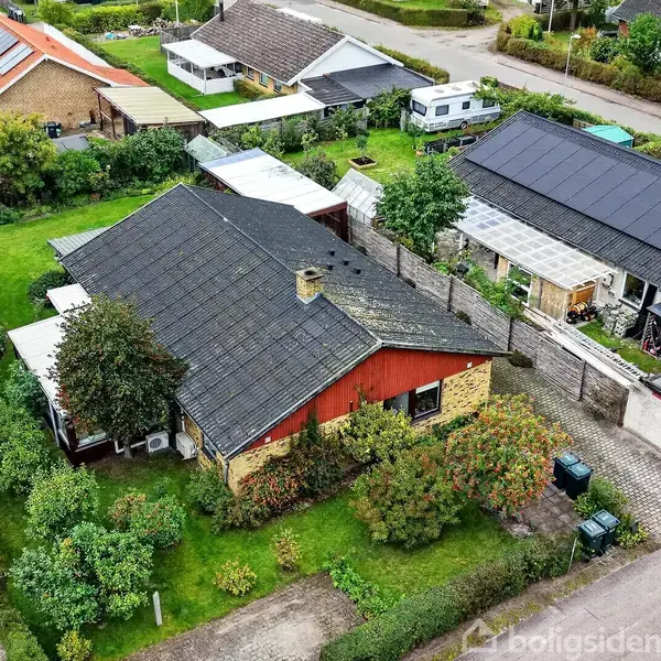 Hus set ovenfra, omgivet af grønne haver i et parcelhuskvarter. Biler parkeret ved siden af husene, græs og buske i forgrunden. Flere huse og en vej i baggrunden.